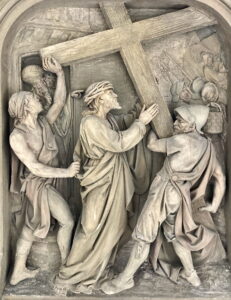 Ein Steinrelief zeigt drei Männer, die einen in ein Gewand gehüllten Mann zwingen, ein großes Holzkreuz zu tragen; die Szene ist detailliert und dramatisch und ruft Emotionen und historische Bedeutung hervor. (Text mit KI erstellt)