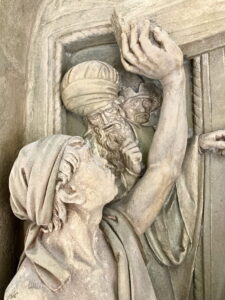 Eine Steinreliefskulptur zeigt eine Person im Vordergrund, die einen Ziegelstein hochhebt, während im Hintergrund zwei Männer in Turbanen und Gewändern zusehen. Die Figuren scheinen mit einer Konstruktion oder einer Bautätigkeit beschäftigt zu sein. (Text mit KI erstellt)