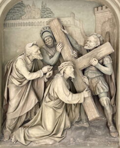 Ein detailliertes Steinrelief zeigt Jesus, der ein großes Kreuz trägt, umgeben von drei Männern, darunter ein römischer Soldat in Rüstung. Ein Mann gestikuliert eindringlich, während ein anderer Jesus stützt, der unter dem Gewicht des Kreuzes kniet. (Text mit KI erstellt)