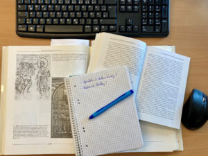 Ein Schreibtisch mit einer Tastatur, einer Maus, zwei aufgeschlagenen Büchern mit Texten und Bildern sowie einem Notizbuch mit handschriftlichen Notizen und einem blauen Stift, das auf den Büchern liegt. (Text mit KI erstellt)