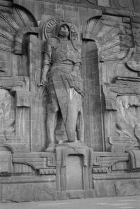Eine große Steinstatue eines Kriegers in Rüstung ist in eine Steinmauer eingelassen, die von Strahlen und dekorativen Schnitzereien umgeben ist. Der Stil ist monumental und detailliert und erinnert an Stärke und Schutz. (Text mit KI erstellt)