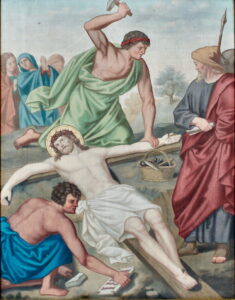 Ein Gemälde zeigt Jesus, der auf einem Holzkreuz liegt, umgeben von Männern, die seine Kreuzigung vorbereiten. Ein Mann hält einen Hammer über die Hand Jesu, ein anderer kniet neben seinen Füßen, und im Hintergrund stehen Schaulustige. (Text mit KI erstellt)