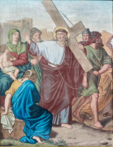 Ein Gemälde zeigt Jesus, der ein großes Holzkreuz trägt, umgeben von römischen Soldaten und trauernden Frauen, von denen eine ein Kind hält, mit einer Stadt im Hintergrund. Jesus trägt einen Heiligenschein, ist in Ketten gelegt und trägt ein Gewand. (Text mit KI erstellt)
