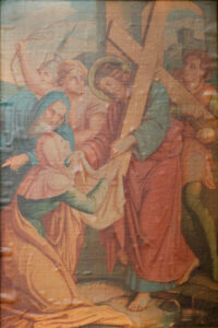 Ein klassisches Gemälde zeigt Jesus, der das Kreuz trägt, umgeben von mehreren Menschen. Eine Frau kniet vor ihm und hält ein Tuch in der Hand, während Jesus nach ihr greift. Im Hintergrund sind Soldaten und Gebäude zu sehen. (Text mit KI erstellt)