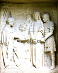 Ein Steinrelief zeigt vier Figuren: einen sitzenden Mann auf der linken Seite, ein Kind, das eine Urkunde hält, und zwei stehende Männer, einer mit einer Dornenkrone, daneben ein römischer Soldat. Drapierte Vorhänge umrahmen den Hintergrund. (Text mit KI erstellt)