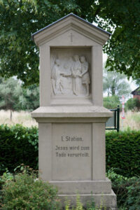 Ein steinernes Denkmal mit einem Relief, das die Verurteilung Jesu zum Tode darstellt, mit der Aufschrift "I. Station. Jesus wird zum Tode verurteilt". Es steht im Freien, umgeben von Grünflächen und Bäumen. (Text mit KI erstellt)