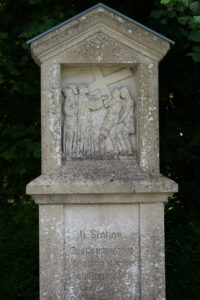 Ein Steinmonument zeigt ein Relief mit drei Figuren, von denen eine ein Kreuz hält. Darunter ist ein verblasster Text eingraviert: "II. Station, Jesus nimmt das Kreuz auf seine Schultern". Im Hintergrund sind Bäume zu sehen. (Text mit KI erstellt)