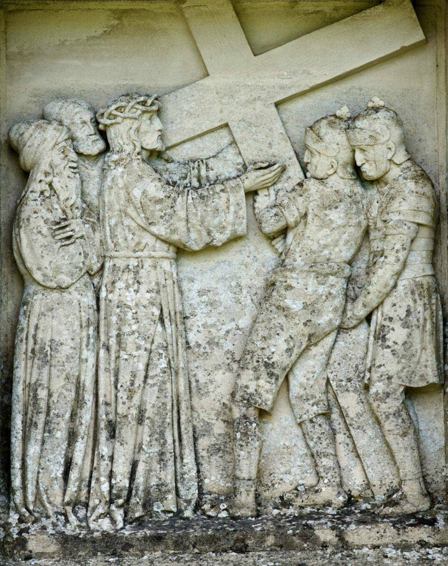 Steinrelief mit der Darstellung von Jesus, der ein Kreuz trägt, flankiert von drei römischen Soldaten und einer Frau, mit verwitterten und flechtenbedeckten Oberflächen. ( )