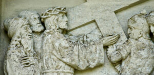 Steinrelief mit der Darstellung von Jesus, der ein Kreuz trägt, mit zwei Männern hinter ihm und einem Soldaten vor ihm, die alle antike Gewänder und Kopfbedeckungen tragen. Die Szene ist mit strukturiertem, verwittertem Stein detailliert dargestellt. (Text mit KI erstellt)