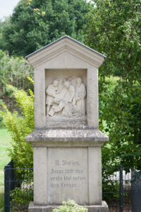 Ein Steindenkmal mit einem Relief, das Jesus unter dem Kreuz fallend darstellt, umgeben von drei Figuren. Die Inschrift darunter lautet: III. Station. Jesus fällt das erste Mal unter dem Kreuze. Im Hintergrund ist grünes Laub zu sehen. (Text mit KI erstellt)