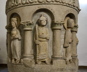 Eine Steinsäule ist mit in Bögen eingearbeiteten Figuren verziert, darunter eine Person mit Helm und Stab, eine Frau, die ein Gefäß hält, sowie eine geflügelte Gestalt. Der obere Rand ist mit dekorativen Mustern und lateinischen Inschriften verziert. (Text mit KI erstellt)
