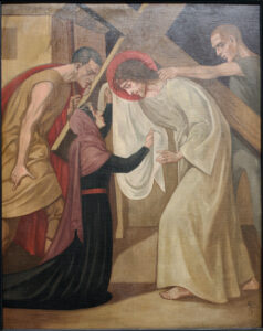 Ein Gemälde zeigt Jesus, wie er ein Kreuz trägt, eine Dornenkrone und ein weißes Gewand trägt und einer knienden Frau begegnet, die sein Gesicht berührt. Zwei Männer stehen daneben, von denen einer das Kreuz hält und der andere die Hand ausstreckt. Die Szene ist düster und bewegend. (Text mit KI erstellt)