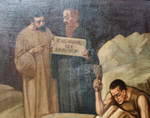 Zwei Männer stehen nebeneinander; einer hält ein Schild mit der Aufschrift „JESUS NAZARENUS REX JUDAEORUM“, während ein dritter Mann darunter mit Hammer und Meißel in Stein meißelt. Der Hintergrund ist dunkel und felsig und erinnert an eine biblische oder historische Szene. (Text mit KI erstellt)