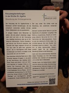 Eine Person hält einen gedruckten deutschen Informationsflyer mit dem Titel „Kreuzwegdarstellungen in der Kirche St. Agatha“ in der Hand, in dem die Darstellung der Kreuzwegstationen und die Haltung der Kirche gegen antisemitische Interpretationen erläutert werden. (Text mit KI erstellt)