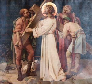 Ein Gemälde zeigt Jesus, wie er ein hölzernes Kreuz trägt, eine Dornenkrone auf dem Kopf und einen leuchtenden Heiligenschein um den Kopf. Um ihn herum stehen drei Männer, die ihm offenbar helfen oder das Kreuz stützen. Die Szene spielt im Freien; auf dem Boden sind Felsen und vereinzelte Pflanzen zu sehen. (Text mit KI erstellt)