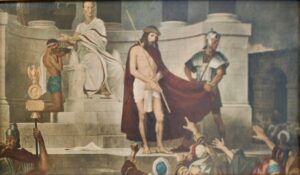 Jesus, der eine Dornenkrone und ein rotes Gewand trägt, steht vor einer Menschenmenge. Ein römischer Beamter gibt von einem thronartigen Sitz aus Gesten, während ein Soldat Jesus festhält. Im Vordergrund strecken Menschen ihre Hände aus, manche flehend, vor dem Hintergrund einer steinernen Architektur. (Text mit KI erstellt)