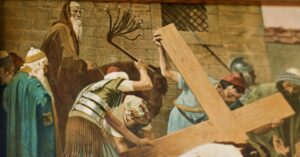 Ein historisches Gemälde zeigt Soldaten, die einen Mann zwingen, ein großes Holzkreuz zu tragen, während Zuschauer in Gewändern zusehen. Im Hintergrund sind eine Steinmauer und ein vergittertes Fenster zu sehen. (Text mit KI erstellt)