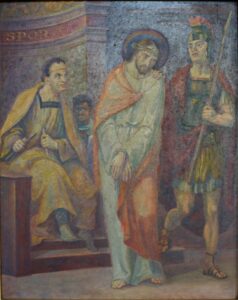 Ein Gemälde zeigt Jesus mit gefesselten Händen, wie er zwischen einem sitzenden römischen Beamten und einem Speer tragenden Wachsoldaten steht. Der Beamte macht eine Geste, und der Wachsoldat trägt eine Rüstung und einen Helm. Im Hintergrund sind Säulen und Vorhänge zu sehen. (Text mit KI erstellt)