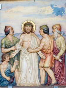 Eine gemalte Darstellung von Jesus mit Heiligenschein, umgeben von vier Männern, von denen einer kniet, während sie ihm das Gewand ausziehen; im Hintergrund ist eine hügelige Landschaft zu sehen. (Text mit KI erstellt)