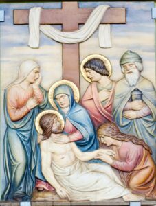 Ein gemaltes Relief zeigt fünf Menschen, die über den Leichnam Jesu trauern, der vom Kreuz herabgenommen wurde. Im Hintergrund ist ein großes Holzkreuz zu sehen, das mit einem weißen Tuch bedeckt ist. Die Figuren tragen goldene Heiligenscheine und zeigen traurige Gesichtsausdrücke. (Text mit KI erstellt)