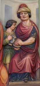 Ein geschnitztes und bemaltes Relief zeigt eine sitzende Person in einem roten Gewand und mit einem Hut, die einen Krug in der Hand hält, während neben ihr eine jüngere Person einen weiteren Krug darreicht; beide haben ernste Gesichtsausdrücke und tragen detailreich gestaltete Kleidung im klassischen Stil. (Text mit KI erstellt)