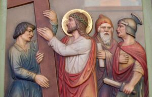 Ein Relief zeigt Jesus, wie er ein Kreuz trägt; um seinen Kopf ist ein Heiligenschein zu sehen. Er wird von drei Männern flankiert – einem in Blau, einem weiteren in Rot mit einem Hut und einem Soldaten in Rüstung –, die jeweils Schriftrollen oder Gegenstände in den Händen halten. (Text mit KI erstellt)