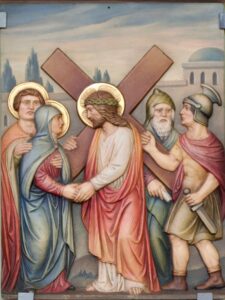 Jesus, der eine Dornenkrone trägt, schleppt ein Kreuz, als er einer Frau begegnet. Neben ihm stehen zwei Gestalten mit Heiligenschein. Zwei Männer, von denen einer ein Schwert hält, schauen zu. Im Hintergrund sind Gebäude und Bäume zu sehen, was auf eine biblische Szene hindeutet. (Text mit KI erstellt)