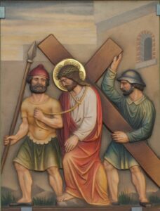 Eine Darstellung von Jesus, der ein Kreuz trägt, flankiert von zwei Soldaten; der eine hält einen Speer, der andere stützt das Kreuz. Jesus trägt einen Heiligenschein und ein rotes Gewand, während die Soldaten Helme und Tuniken tragen. Die Szene scheint auf eine Wand gemalt zu sein. (Text mit KI erstellt)