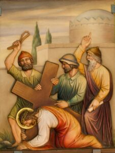 Religiöses Gemälde, das Jesus zeigt, wie er kniend ein hölzernes Kreuz trägt, mit einem Heiligenschein um den Kopf. Dahinter stehen drei Männer in Gewändern: Einer hält eine Peitsche, ein anderer deutet nach oben, und der dritte stützt das Kreuz. Im Hintergrund sind eine Steinmauer und Bäume zu sehen. (Text mit KI erstellt)