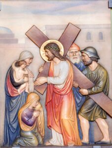 Ein Relief zeigt Jesus, wie er ein Kreuz trägt, eine Dornenkrone auf dem Kopf hat und von Trauernden und einem Soldaten umgeben ist. Eine Frau kniet, eine andere hält ein Kind im Arm, und hinter ihnen steht ein älterer Mann. Jesus hat einen Heiligenschein, und im Hintergrund sind Steingebäude zu sehen. (Text mit KI erstellt)