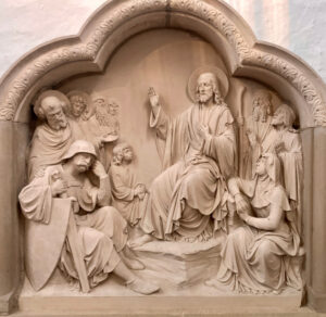 Steinrelief, das eine sitzende zentrale Figur mit erhobener Hand darstellt, umgeben von aufmerksamen Begleitern in verschiedenen Posen – teils sitzend, teils stehend –, in einem detailreichen, gewölbten Rahmen. (Text mit KI erstellt)