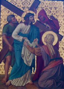 Jesus trägt ein Kreuz und eine Dornenkrone; er wird von drei weiteren Personen begleitet, von denen eine kniet und ihm ein Tuch reicht. Alle sind mit Heiligenscheinen dargestellt. Der Hintergrund besteht aus einem goldenen, kunstvollen Muster. (Text mit KI erstellt)