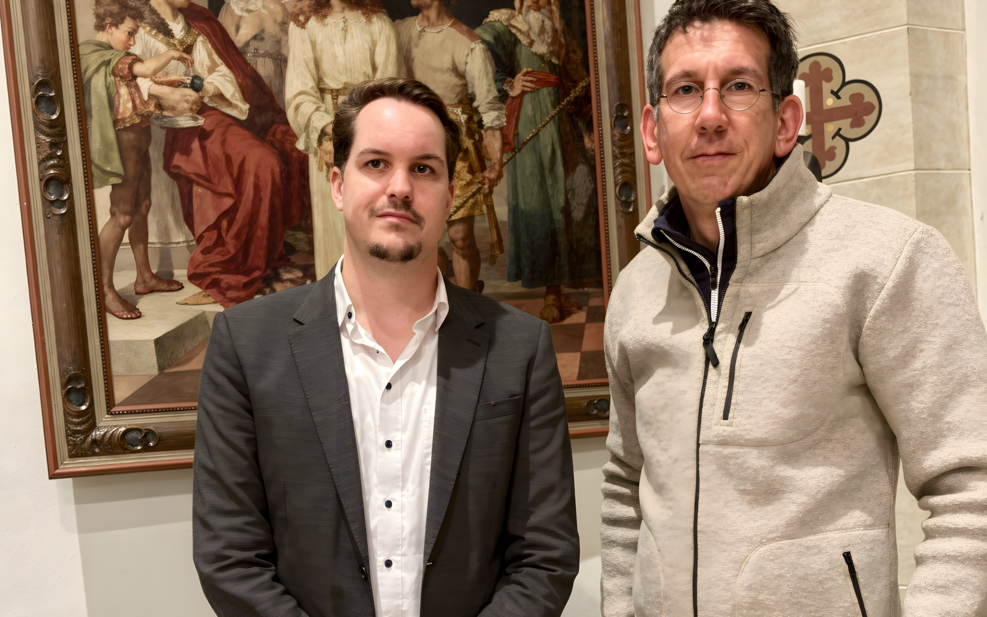 Zwei Männer stehen in einem Innenraum vor einem großen, gerahmten Gemälde, das biblische oder historische Figuren zeigt. Der eine Mann trägt einen dunklen Anzug und ein weißes Hemd, der andere eine Brille und eine helle Jacke. Die Umgebung wirkt formell, möglicherweise handelt es sich um eine Galerie oder ein Museum. (Text mit KI erstellt)